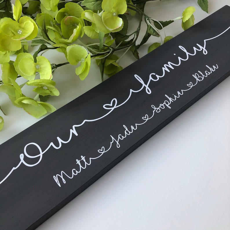 May include: A black wooden sign with the words "Our family" in white script. Below the word "family" are the names Matt, Jadu, Sophie, and Blake, also in white script. The sign is decorated with a white heart design.