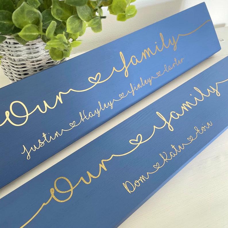 May include: Two blue wooden signs with gold lettering. The signs read "Our Family" with names below. The names are connected by a gold line with hearts. A green plant in a white basket is in the background.