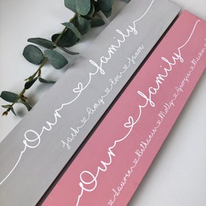 May include: Two wooden family signs, one gray and one pink, with white script. The gray sign reads "Our family" with names Jack, Amy, and Evax. The pink sign reads "Our family" with names Lauren, Bethanie, Molly, and Maisie.
