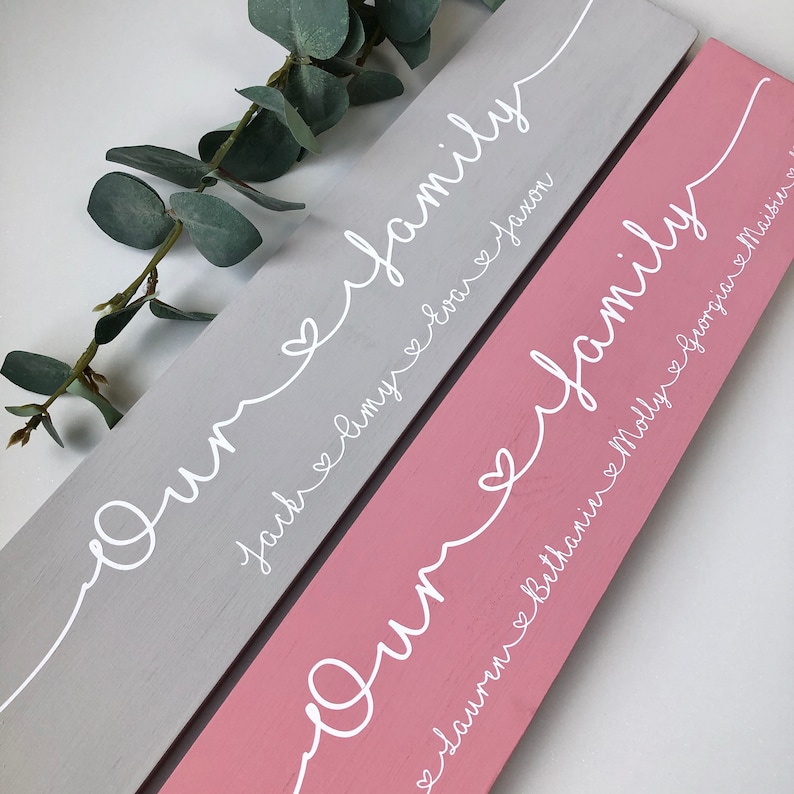 May include: Two wooden family signs, one gray and one pink, with white script. The gray sign reads "Our family" with names Jack, Amy, and Evax. The pink sign reads "Our family" with names Lauren, Bethanie, Molly, and Maisie.
