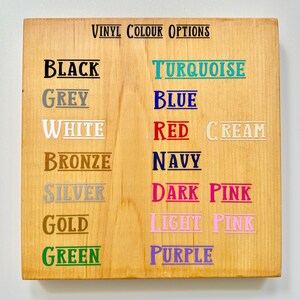 May include: A wooden sign displaying "VINYL COLOUR OPTIONS" in black text. Below, various color names are listed, including black, grey, white, bronze, silver, gold, and green. Additional colors are turquoise, blue, red, cream, navy, dark pink, light pink, and purple.