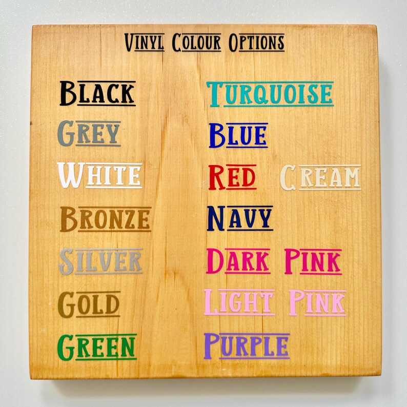 May include: A wooden sign displaying "VINYL COLOUR OPTIONS" in black text. Below, various color names are listed, including black, grey, white, bronze, silver, gold, and green. Additional colors are turquoise, blue, red, cream, navy, dark pink, light pink, and purple.