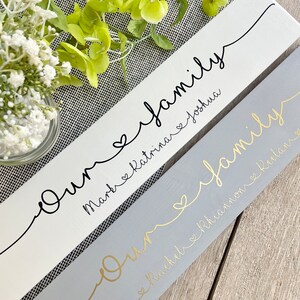 May include: Two wooden signs with the word "Our family" in cursive script. One sign is white with black text, the other is gray with gold text. Names are written below the word "family". A heart symbol is between the names.