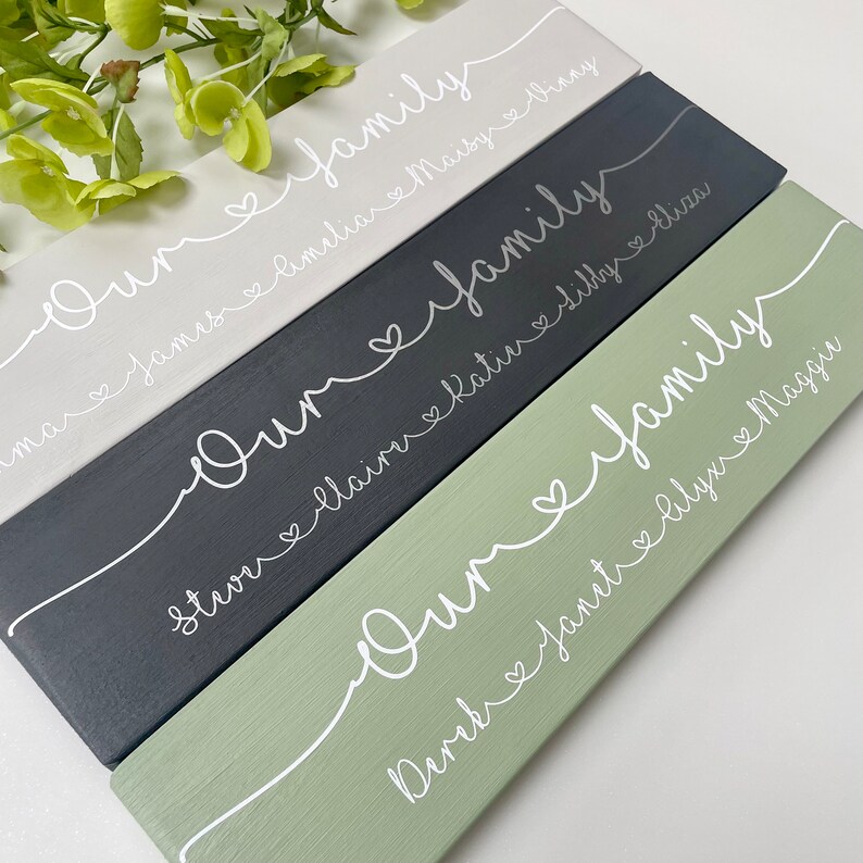 May include: Four rectangular wooden signs in shades of gray, black, and green. Each sign has the words "Our family" in white script, with names and hearts below. Decorative home decor.