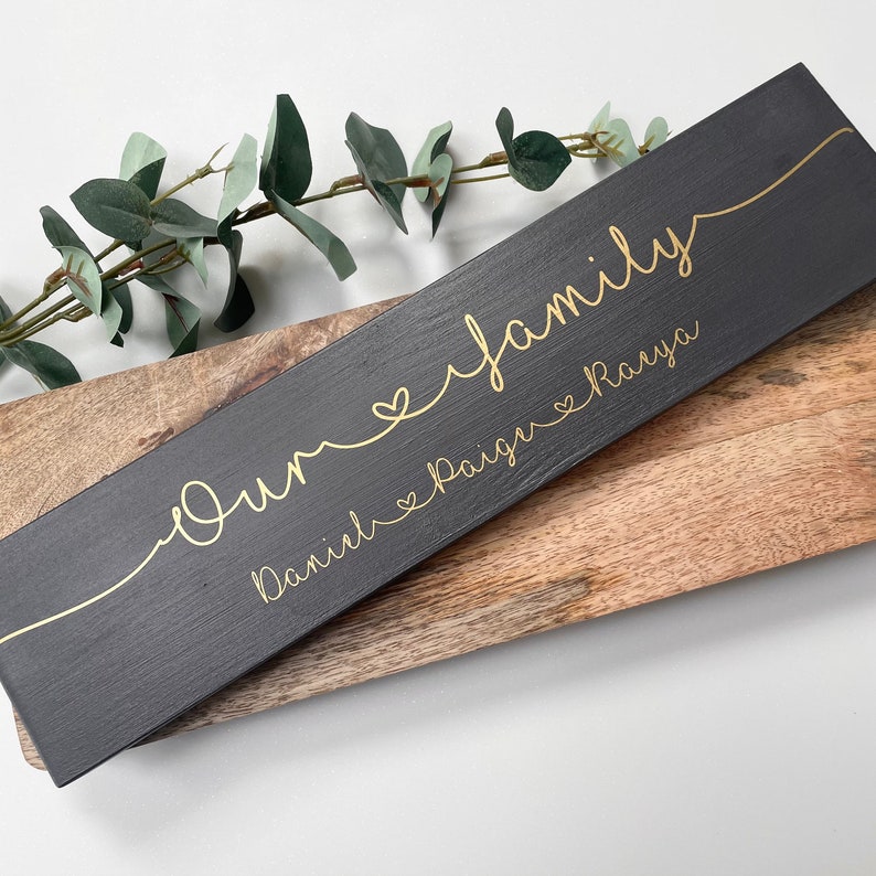 May include: A rectangular, dark gray wooden sign with gold lettering that reads "Our family" in cursive, with the names "Daniel, Paige, and Raya" below. The sign is placed on a wooden surface with a sprig of greenery.