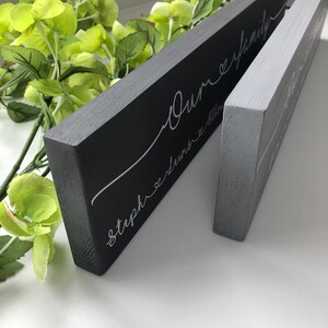 May include: Two rectangular wooden signs, one dark gray and one light gray, with white cursive text. The larger sign reads "Our family" with names below. The smaller sign has similar text. Green foliage is in the background.