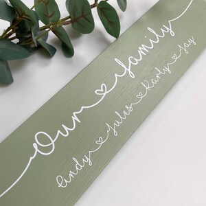 May include: A sage green wooden sign with the words "Our family" in white script. Below, the names "Lindy, Julius, Karly, Jay" are also written in white script. Each name is connected by a heart symbol.
