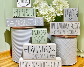 May include: A collection of decorative wooden laundry room signs in various shapes and sizes. The signs feature phrases like "Laundry," "Self Service," and "Wash, Dry, Fold, Repeat." The color palette includes white, gray, and light green with black lettering.
