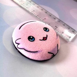 Puede incluir: Un peluche rosa en forma ovalada con una cara sonriente, ojos azules y contornos negros. El peluche tiene un borde negro y está junto a una regla transparente. El peluche mide aproximadamente 5 cm de largo.