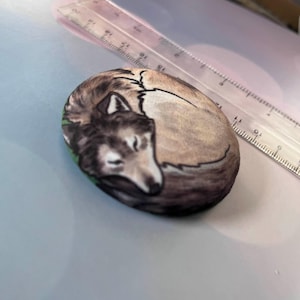 Puede incluir: Una piedra ovalada pintada a mano representa un lobo durmiente. El lobo está representado en tonos marrones, grises y blancos, acurrucado sobre un parche verde. La piedra mide aproximadamente 10 cm de largo.