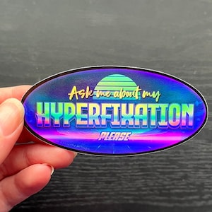 May include: Oval sticker with a holographic finish. The sticker has a retro sunset design and the text "Ask me about my hyperfixation please".