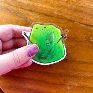 Puede incluir: Una pegatina de monstruo de slime verde con una cara sonriente y varios objetos en su interior, incluyendo una espada, un escudo y huesos. La pegatina está sobre un fondo transparente.