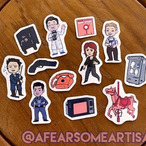 May include: A set of 10 stickers featuring various illustrations, including a safe, a gun, a rotary phone, a television, a carousel horse, a woman with red hair, and several characters. The stickers are on a wooden surface.