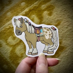May include: A brown and white spotted horse with a brown saddle and a floral crown. The horse is standing on a white background.