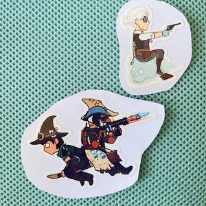 以下が含まれることがあります： 漫画のキャラクターが描かれた2つのダイカットステッカー。1つは、頭蓋骨に座っている銃を持った人物を示しています。もう1つは、魔女の帽子とライフルを持った2人のキャラクターを描いています。ステッカーは白い縁取りで、緑色のメッシュの背景にあります。