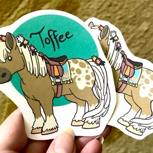 May include: A sticker of a brown and white horse with a saddle and a floral crown. The horse is standing on a teal background with the word "Toffee" written in black.