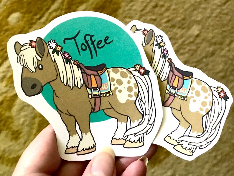 May include: A sticker of a brown and white horse with a saddle and a floral crown. The horse is standing on a teal background with the word "Toffee" written in black.