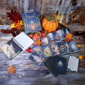 May include: A Gnostic Codes oracle deck set featuring a guidebook, a box with a temple illustration, and numerous cards with celestial and ethereal imagery. An orange pumpkin, a lit candle, and autumn leaves are arranged around the items.