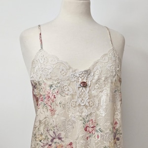 May include: Cream-coloured slip dress featuring a floral pattern in pink, purple, and green. The neckline and straps have lace trim. A small rose adorns the bodice. The dress is displayed on a mannequin, showcasing its delicate design and vintage style.