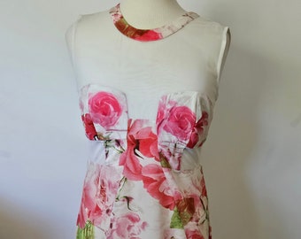 May include: A sleeveless white dress with a floral rose pattern in shades of pink and red. The dress has a sheer neckline and bodice overlay. The roses are large and detailed, with green leaves. The dress is displayed on a mannequin.