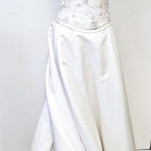 May include: A floor-length white gown featuring a fitted bodice with delicate silver floral embroidery and beadwork, thin spaghetti straps, and a smooth, flowing skirt. The dress is displayed on a mannequin with a matching beaded choker.