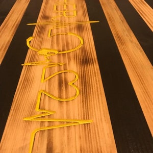 K&ouml;nnte beinhalten: Eine amerikanische Flagge aus Holz mit schwarzen und gelben Streifen. Die Flagge hat eine gelbe Inschrift mit der Aufschrift "523".