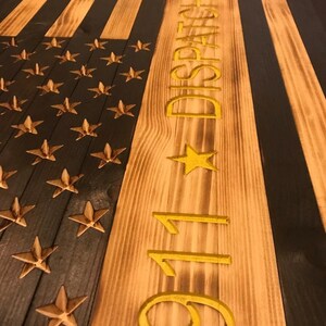 K&ouml;nnte beinhalten: Eine amerikanische Holzflagge mit schwarzen und braunen Streifen und goldenen Sternen. Die Flagge tr&auml;gt die Aufschrift "911 DISPATCHER" in Gold auf der rechten Seite.