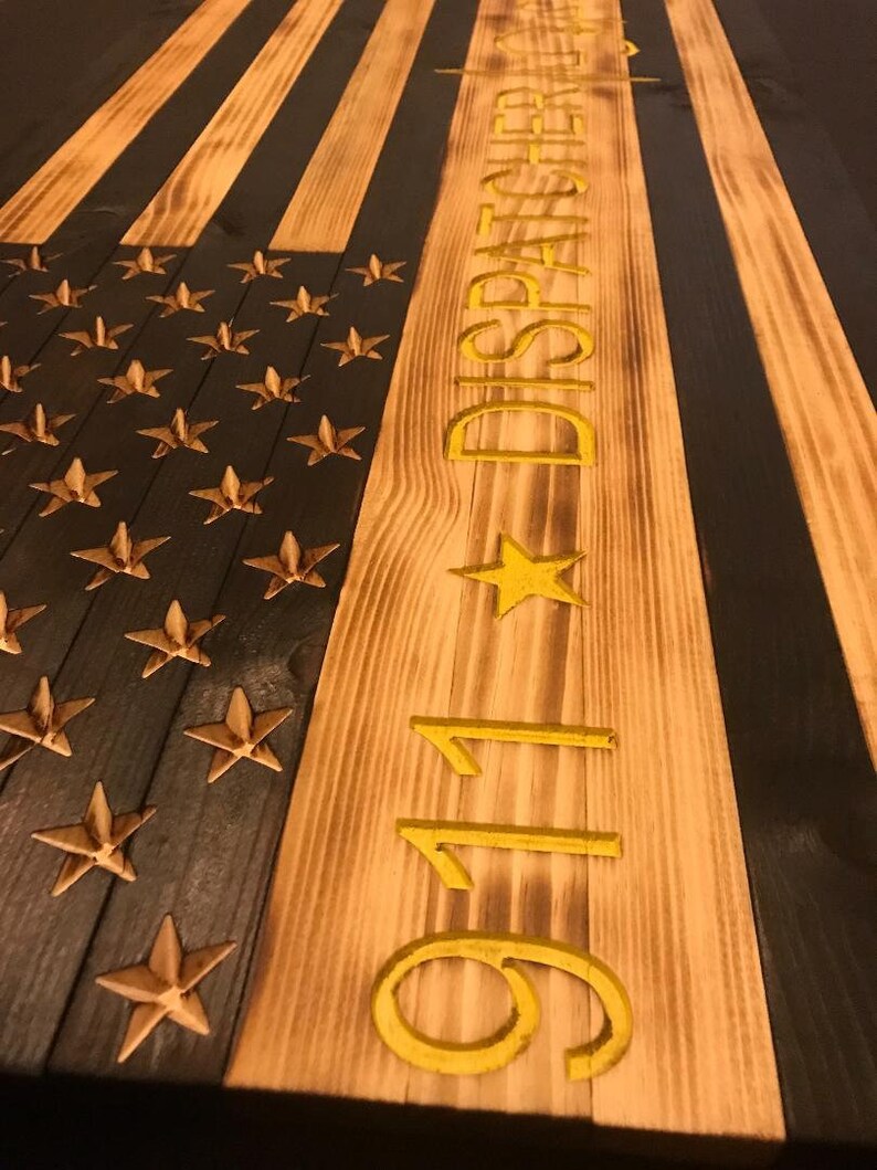 K&ouml;nnte beinhalten: Eine amerikanische Holzflagge mit schwarzen und braunen Streifen und goldenen Sternen. Die Flagge tr&auml;gt die Aufschrift "911 DISPATCHER" in Gold auf der rechten Seite.