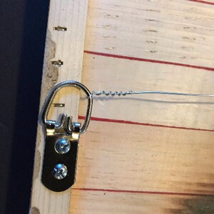 K&ouml;nnte beinhalten: Ein silberner Metall-Haken zum Aufh&auml;ngen mit zwei Schrauben, die an einem Holzrahmen befestigt sind. Der Haken ist an einem Draht befestigt.