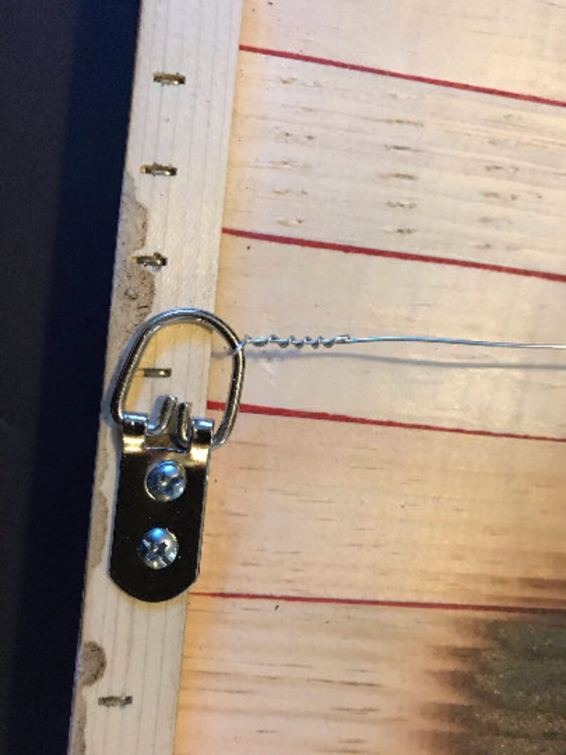 K&ouml;nnte beinhalten: Ein silberner Metall-Haken zum Aufh&auml;ngen mit zwei Schrauben, die an einem Holzrahmen befestigt sind. Der Haken ist an einem Draht befestigt.