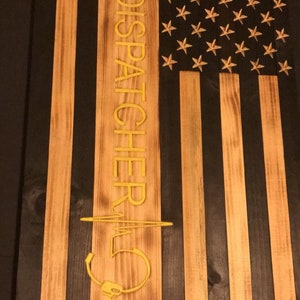 K&ouml;nnte beinhalten: Eine amerikanische Holzflagge mit schwarzem Hintergrund und goldenen Sternen. Die Flagge hat einen vertikalen Streifen mit dem Text "911 DISPATCHER" und einem stilisierten Herzschlag mit einem Headset.