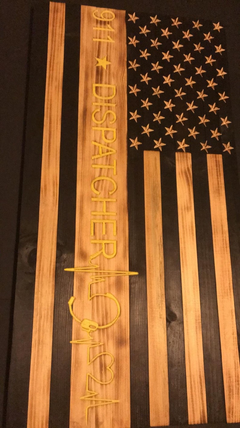 K&ouml;nnte beinhalten: Eine amerikanische Holzflagge mit schwarzem Hintergrund und goldenen Sternen. Die Flagge hat einen vertikalen Streifen mit dem Text "911 DISPATCHER" und einem stilisierten Herzschlag mit einem Headset.