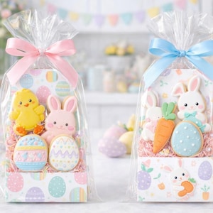 Könnte beinhalten: Zwei Geschenkbeutel mit Osterkeksen. Einer hat eine rosa Schleife und zeigt ein gelbes Küken, einen rosa Hasen und dekorierte Eier. Der andere Beutel hat eine blaue Schleife und enthält Hasenkekse, eine Karotte und ein Ei. Die Beutel sind in Klarsichtfolie verpackt.