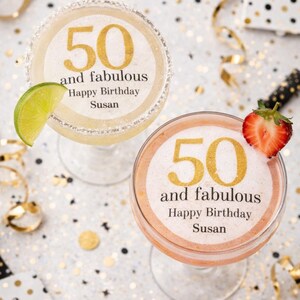 May include: Two margarita glasses with the text "50 and fabulous Happy Birthday Susan" in gold. One glass has a lime wedge, the other a strawberry. The background is white with gold confetti and ribbons.