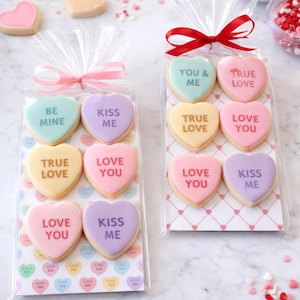 Könnte beinhalten: Herzförmige Kekse in Zellophantüten, die mit Bändern gebunden sind. Die Kekse sind in Pastellfarben gehalten und tragen Aufschriften wie "Be Mine" und "Love You". Streusel und lose herzförmige Süßigkeiten sind verstreut.