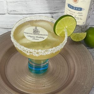 May include: A margarita cocktail in a large glass with a salt-rimmed edge, garnished with a lime slice. The glass sits on a gold-colored plate. The cocktail has a label that reads "TEQUILA MOCKINGBIRD Happy Birthday Susan". A bottle of Tequila Ocho is in the background.