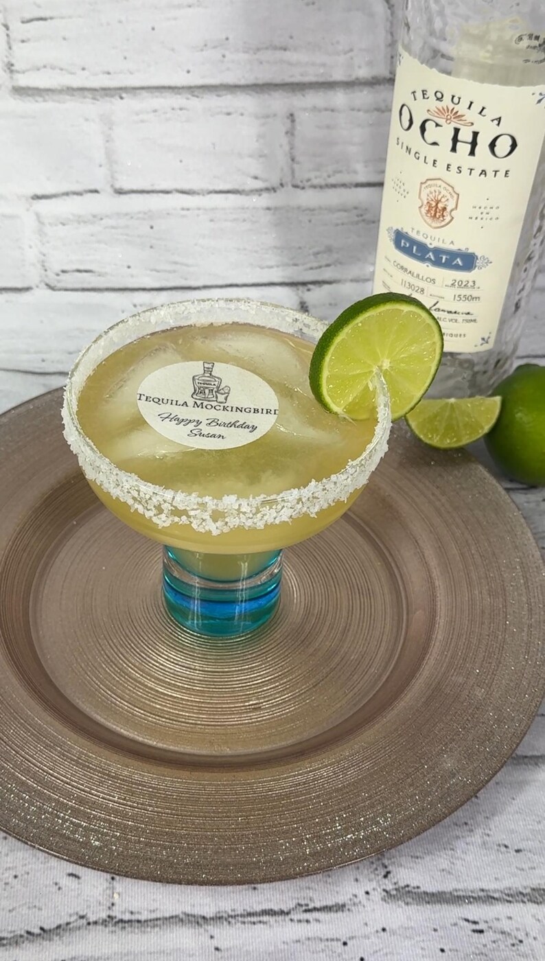 May include: A margarita cocktail in a large glass with a salt-rimmed edge, garnished with a lime slice. The glass sits on a gold-colored plate. The cocktail has a label that reads "TEQUILA MOCKINGBIRD Happy Birthday Susan". A bottle of Tequila Ocho is in the background.
