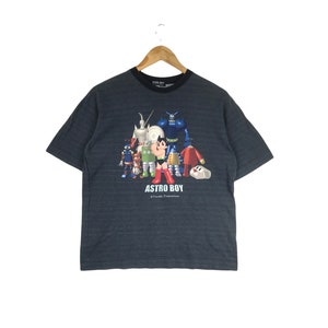 May include: A dark blue t-shirt featuring a graphic print of Astro Boy and other characters from the Astro Boy anime series. The text "ASTRO BOY" is printed below the graphic. The text "© Tezuka Productions" is printed below the text "ASTRO BOY".