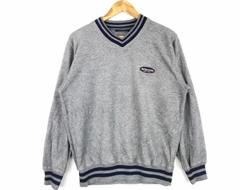 May include: A gray V-neck sweatshirt with blue and white stripes on the collar and cuffs. The sweatshirt has a small oval logo on the front that says "Vans Off The Wall".