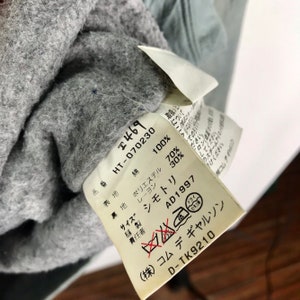 Op de afbeelding: Een wit label met zwarte tekst die de samenstelling van een kledingstuk beschrijft. Het label luidt "100% katoen", "70% polyester", "30% rayon", "Maat", "Naaiwerk", "Verantwoordelijke", "AD1997", "(株)コムデギャルソン", "D-TK9210".