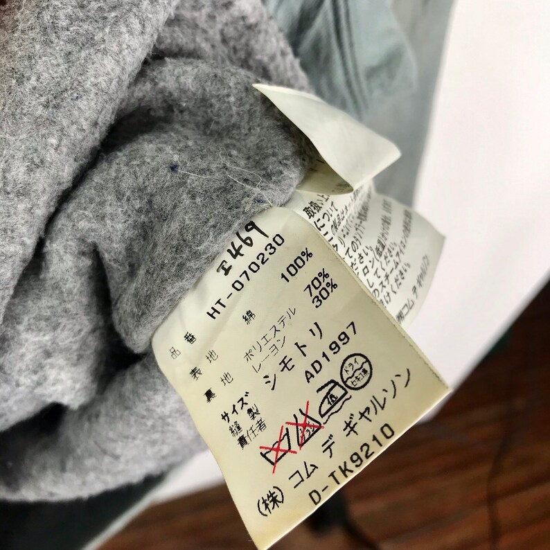 Op de afbeelding: Een wit label met zwarte tekst die de samenstelling van een kledingstuk beschrijft. Het label luidt "100% katoen", "70% polyester", "30% rayon", "Maat", "Naaiwerk", "Verantwoordelijke", "AD1997", "(株)コムデギャルソン", "D-TK9210".