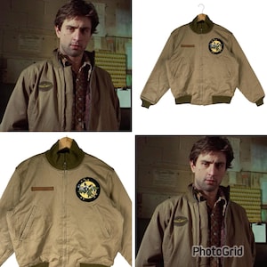 May include: A tan zip-up bomber jacket featuring a dark olive green ribbed collar, cuffs, and hem. The jacket includes a circular patch on the left chest with a graphic of a creature and stars, and a rectangular patch on the right chest area.