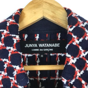 May include: Close-up of a navy blue jacket with a red and white geometric pattern. The label reads "JUNYA WATANABE COMME des GAR&Ccedil;ONS" with a size "M". The jacket is hanging on a wooden hanger.