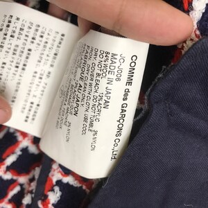 May include: A close-up of a tag from a Comme des Gar&ccedil;ons garment. The tag reads "MADE IN JAPAN" and lists the fabric composition: 84% wool, 13% acrylic, and 3% nylon. The brand name is also visible.