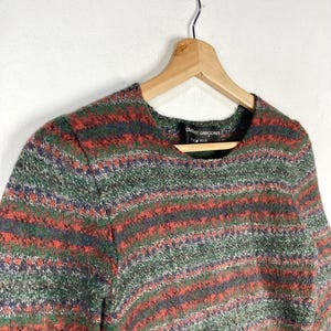 May include: A striped sweater with a green, red, and blue color scheme. The sweater has a round neckline and long sleeves. The label on the sweater reads "Comme des Gar&ccedil;ons" and "Homme Plus".