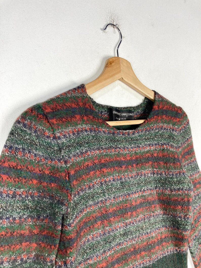 May include: A striped sweater with a green, red, and blue color scheme. The sweater has a round neckline and long sleeves. The label on the sweater reads "Comme des Gar&ccedil;ons" and "Homme Plus".