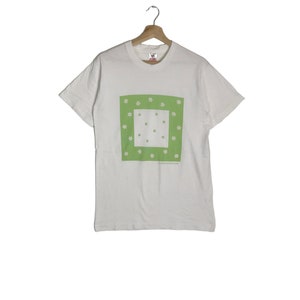 May include: White t-shirt with a green square design featuring white flowers. The shirt is hanging on a wooden hanger. The text on the shirt is small and difficult to read.