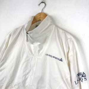 May include: White windbreaker jacket with a blue embroidered logo that says "Le Coq Sportif" on the left side and a blue embroidered logo that says "Le Coq Sportif" on the right side.