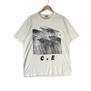 May include: Cream-colored t-shirt with a black graphic print of a building and vertical lines. The letters "C.E" are printed in black below the graphic. The t-shirt is hanging on a wooden hanger.
