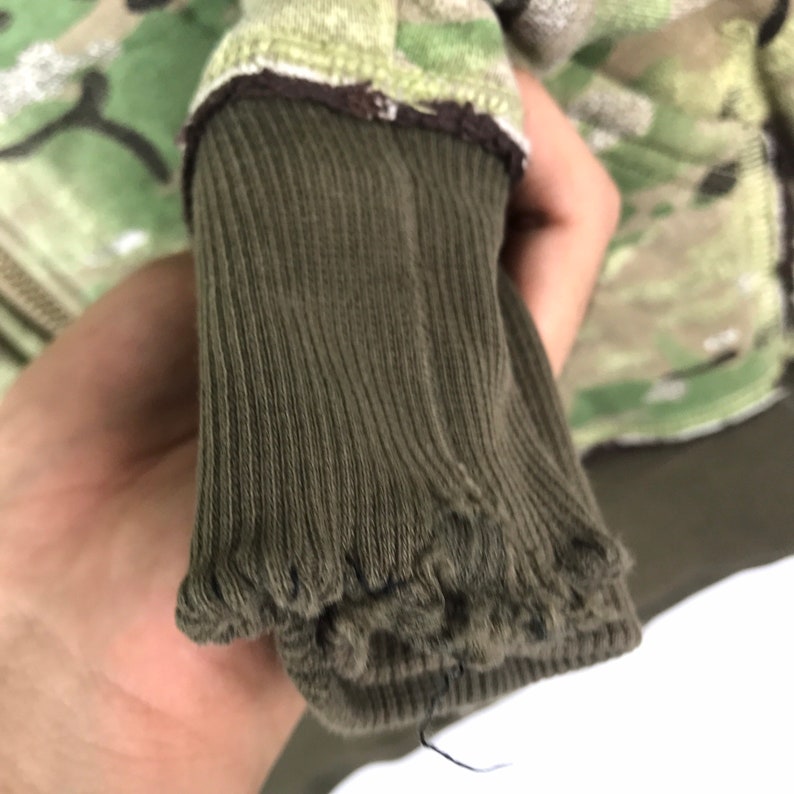 May include: Close-up of a green camouflage jacket with a brown ribbed cuff. The cuff is folded over and shows the stitching.